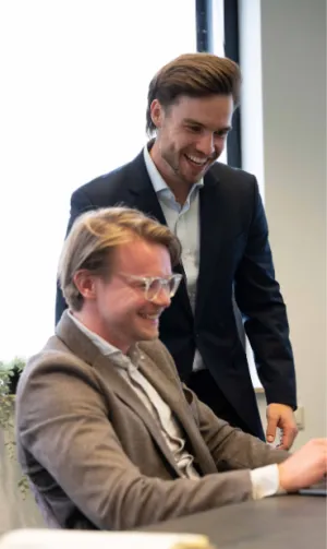 Twee mannen in pak lachen samen terwijl ze naar een computerscherm kijken op een kantoor.