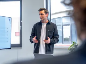  Jonge man geeft een presentatie in een modern kantoor, staand voor een raam en gekleed in een zwarte overshirt en wit T-shirt.