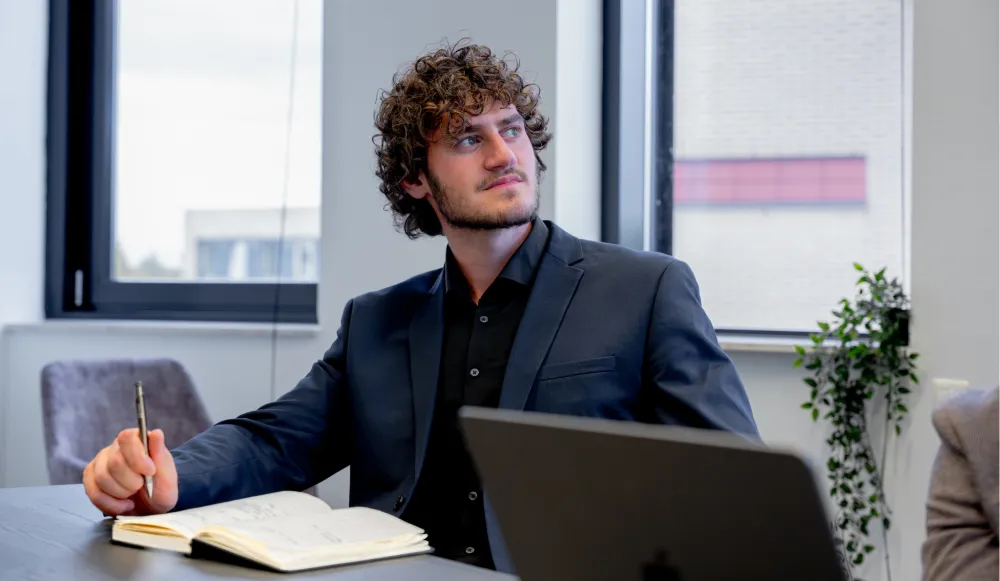 Man in donker pak zit aan een vergadertafel met een open notitieboek en kijkt nadenkend uit het raam.