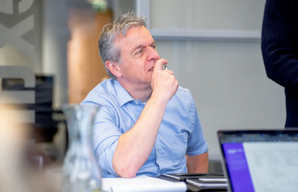 Man in lichtblauw overhemd zit aan een vergadertafel en kijkt nadenkend voor zich uit met zijn hand bij zijn mond.