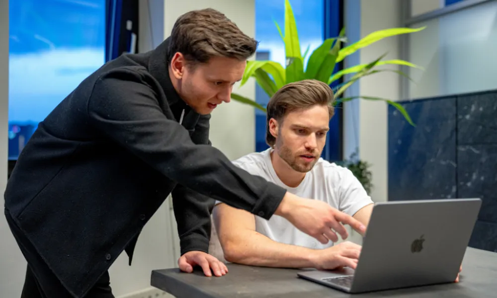 Twee jonge mannen werken samen aan een laptop; de een zit en kijkt geconcentreerd, terwijl de ander staat en iets aanwijst op het scherm.