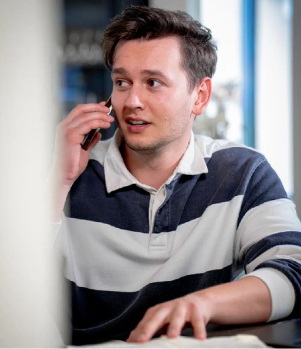 Jongeman in een gestreepte trui voert een telefoongesprek terwijl hij aan een tafel zit.